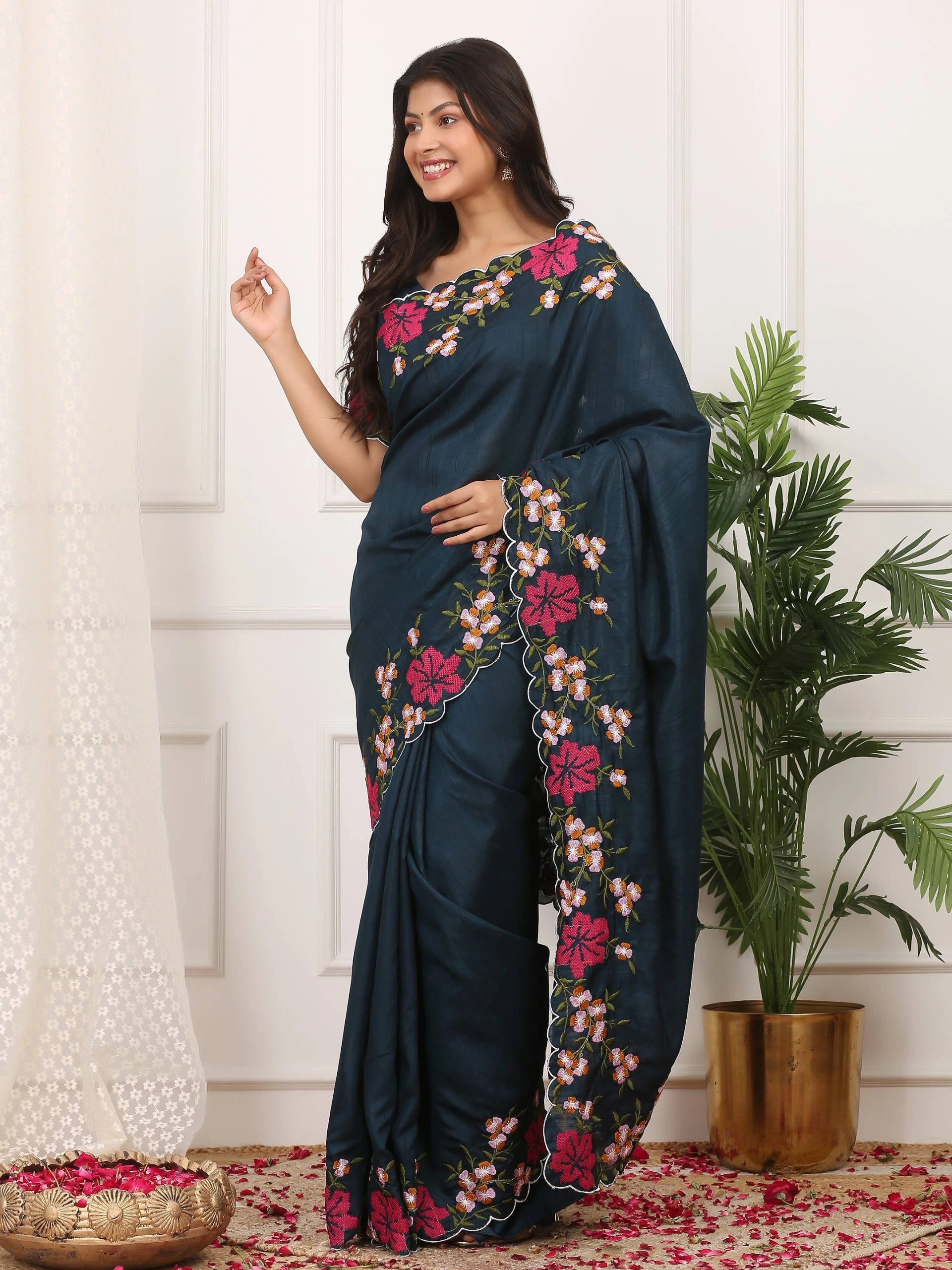 Woman wearing a dark blue saree with floral patterns in a room with white walls and a plant.